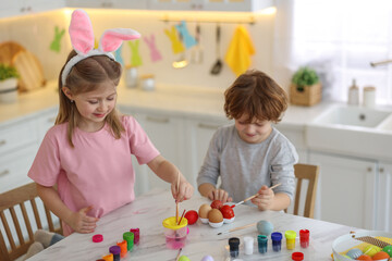 Wall Mural - Easter celebration. Cute children with bunny ears painting eggs at white marble table in kitchen