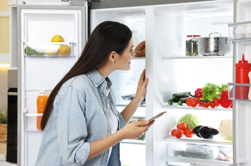 Wall Mural - Young woman with smartphone near modern refrigerator in kitchen