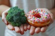 A person holding broccoli in one hand and a pink donut with sprinkles on the other