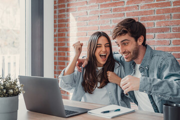 overjoyed millennial couple sit at desk look at laptop screen feel euphoric with online win, excited