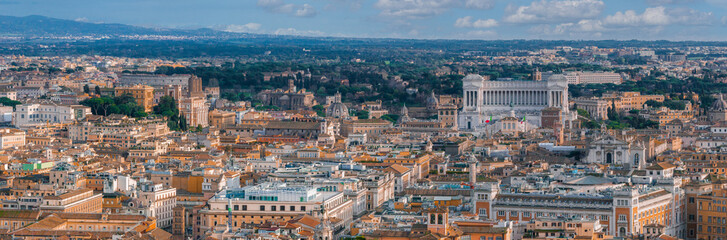 Panoramic aerial view of Rome, Italy, shows terracotta rooftops and landmarks like the Altare della Patria. The city blends with hills under a partly cloudy sky.