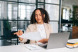 Focused busy curly brazilian or latino woman in elegant clothes, office employee, secretary or financial manager, sits at her workplace with laptop in the modern office, studying documents, analyzing