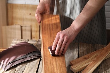 Man polishing wooden plank with sandpaper at table indoors, closeup