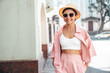 Young beautiful smiling hipster woman in trendy summer pink costume clothes. Carefree female posing in the street at sunny day. Positive model outdoors at sunset. Cheerful and happy in hat, sunglasses