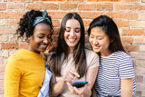 Fototapeta Londyn - Happy young girls using mobile phone standing over brick wall background outdoors. Social media concept.