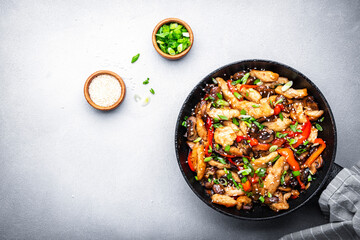 Wall Mural - Asian stir fry chicken  slices with red paprika, mushrooms, chives and sesame seeds in frying pan. Gray kitchen table background, top view