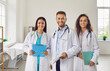 © Studio Romantic - Successful team of medical doctors are looking at camera and smiling while standing in hospital. Portrait of three young medical colleagues in white coats wearing stethoscopes and holding clipboards.