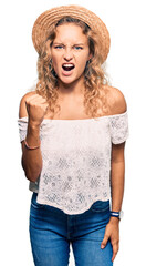 Wall Mural - Beautiful caucasian woman wearing summer hat angry and mad raising fist frustrated and furious while shouting with anger. rage and aggressive concept.