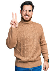 Wall Mural - Young hispanic man wearing casual clothes smiling with happy face winking at the camera doing victory sign with fingers. number two.