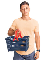 Wall Mural - Young handsome hispanic man holding supermarket shopping basket thinking attitude and sober expression looking self confident