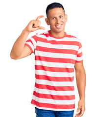 Wall Mural - Young handsome latin man wearing casual clothes smiling and confident gesturing with hand doing small size sign with fingers looking and the camera. measure concept.