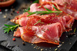 Slices of tasty cured or long-ripened ham with elements of green rosemary and spices close-up on dark slate
