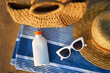 Sunscreen bottle on striped towel next to woven bag, straw hat, and white sunglasses on sandy beach suggests smart sun safety habits while soaking up the seaside ambience on a sunny day.