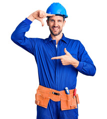 Wall Mural - Young handsome man wearing worker uniform and hardhat smiling making frame with hands and fingers with happy face. creativity and photography concept.