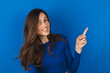 Look, advertise here! Brunette haired pretty woman with appealing smile gives positive attitude towards good deal, expresses her recommendation while pointing on left side isolated on blue background.