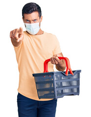 Canvas Print - Young hispanic man wearing shopping basket and medical mask pointing with finger to the camera and to you, confident gesture looking serious