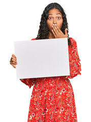 Canvas Print - Young african american girl holding blank empty banner covering mouth with hand, shocked and afraid for mistake. surprised expression