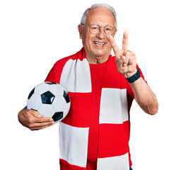 Wall Mural - Senior man with grey hair football hooligan holding ball smiling with happy face winking at the camera doing victory sign with fingers. number two.