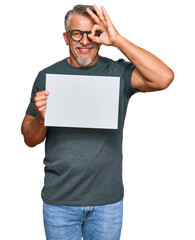 Canvas Print - Middle age grey-haired man holding blank empty banner smiling happy doing ok sign with hand on eye looking through fingers