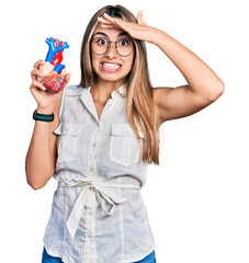 Poster - Hispanic young woman holding heart organ stressed and frustrated with hand on head, surprised and angry face