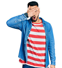 Wall Mural - Young hispanic man with beard wearing casual denim jacket peeking in shock covering face and eyes with hand, looking through fingers with embarrassed expression.