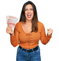 Beautiful hispanic woman holding 10 colombian pesos banknotes screaming proud, celebrating victory and success very excited with raised arms