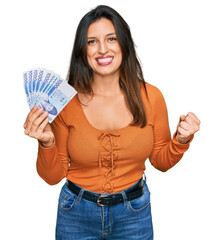 Poster - Beautiful hispanic woman holding 1000 south korean won banknotes screaming proud, celebrating victory and success very excited with raised arms