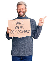 Wall Mural - Young blond man holding save our democracy cardboard banner smiling happy pointing with hand and finger to the side