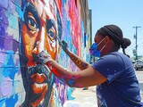 Fototapeta  - A woman is painting a portrait of a man on a wall. The painting is colorful and vibrant, with a mix of blues, reds, and yellows. The woman is wearing a mask