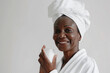 Skincare, cheerful mature African American woman smiling, applying moisturizing cream on face, lotion or mask for skin lifting and anti-aging, wears wrapped towel on head, with plastic jar in hand. 