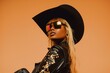 young black woman with blond hair wearing holographic sunglasses black leather cowboy hat and black leather cowboy clothes with a bit of golden accents against a pastel peach backdrop, cowboy core