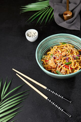 Canvas Print - Chinese fried noodles with chicken and vegetables in ceramic bowl on black concrete background. Modern composition with asian food - homemade noodles in chinese style. Fried udon on wok with sesame.