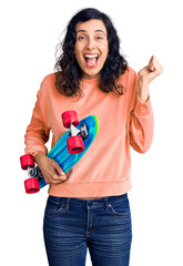 Wall Mural - Young beautiful hispanic woman holding skate screaming proud, celebrating victory and success very excited with raised arms