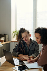 Poster - Two young intercultural businesswomen looking at laptop screen while one of them making notes in copybook during discussion of online data