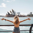 From behind, you can see the traveler girl arms spread wide as she take in the incredible view of the Sydney Opera House in Austailia.