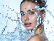 A beautiful woman has water splashing on her face against a white background in a simple composition portraying feminine beauty