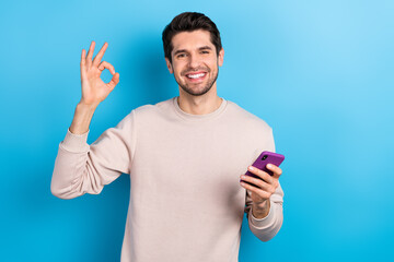 Aufkleber - Photo of good mood cheerful man with stylish haircut dressed beige pullover hold smartphone showing okey isolated on blue color background