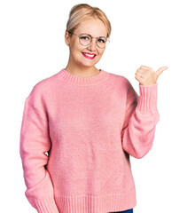 Wall Mural - Young blonde woman wearing casual clothes and glasses smiling with happy face looking and pointing to the side with thumb up.