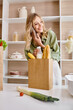 A woman in an apartment kitchen talking on a cell phone while holding a shopping bag.