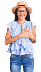 Wall Mural - Young beautiful latin girl wearing summer hat and glasses smiling with hands on chest with closed eyes and grateful gesture on face. health concept.