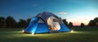 Outdoor camping tent with tarp or flysheet on grass courtyard and warm night light under dark blue sky twilight time