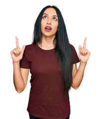 Wall Mural - Young hispanic girl wearing casual t shirt amazed and surprised looking up and pointing with fingers and raised arms.