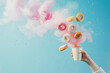 A young woman's hand holds a paper cup into which baby donuts fall from above with colored smoke, light blue background, realistic texture, the donuts are covered with pink and yellow chocolate