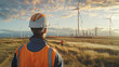 A man in an orange vest stands in a field of wind turbines. The sky is cloudy and the sun is setting