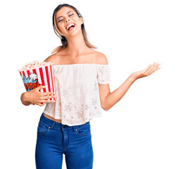 Poster - Young beautiful woman holding popcorn celebrating victory with happy smile and winner expression with raised hands