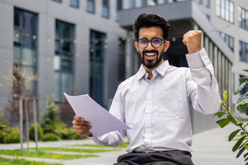 Portrait of a happy Indian male businessman smiling at the camera and happy reading a letter and documents he is holding in his hands showing a victory gesture.