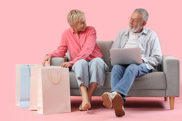 Poster - Mature couple with laptop and shopping bags sitting on sofa against pink background