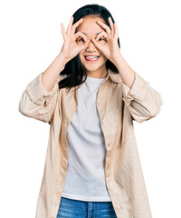 Wall Mural - Young chinese woman wearing casual white t shirt and jacket doing ok gesture like binoculars sticking tongue out, eyes looking through fingers. crazy expression.