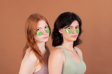 Two young women with under-eye patches posing together on a beige background.