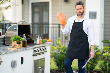 Poster - Man in cook apron cooking salmon fish on barbecue in the backyard of the house. Handsome man preparing salmon barbecue. Barbecue chef master. Grill and barbeque.